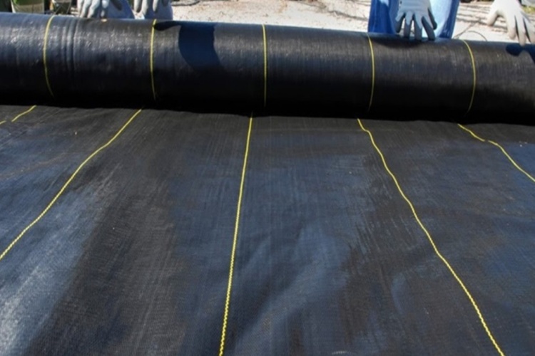 A large roll of black woven geotextile being unrolled at a road construction site