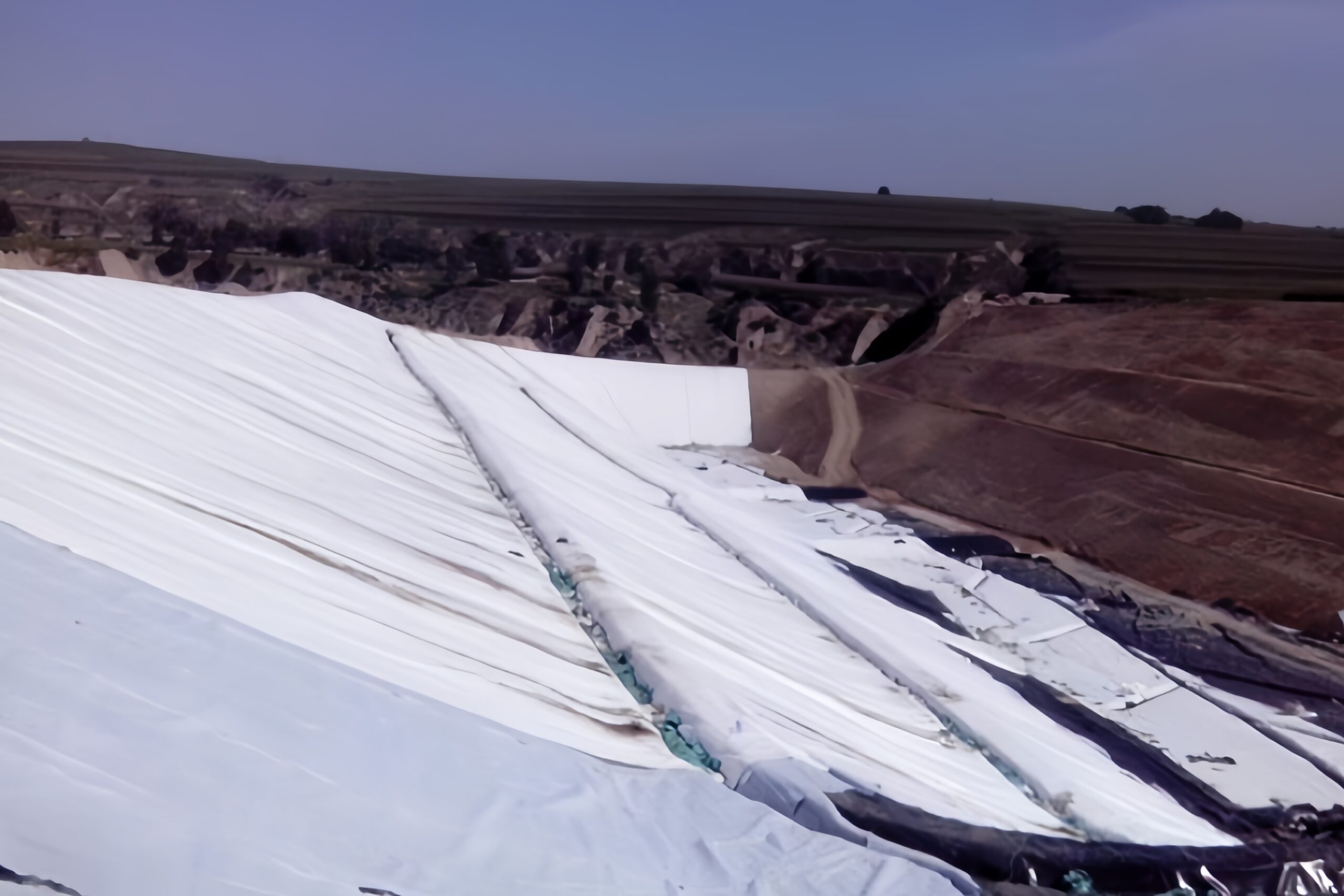 Workers carefully unrolling a geotextile down a long slope, with the top secured in an anchor trench
