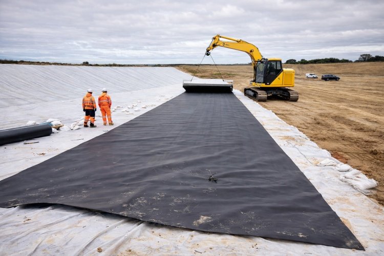 A construction crew deploying a thick, white nonwoven geotextile over a black geomembrane liner for protection.