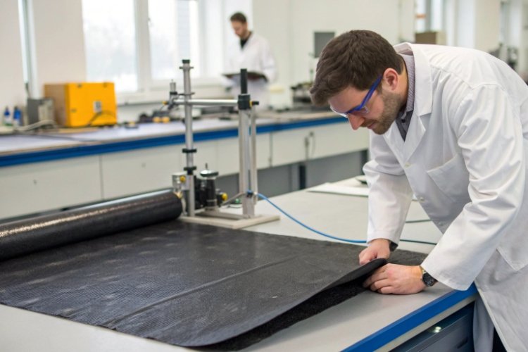 A lab machine performing a tensile strength test on a strip of black geomembrane